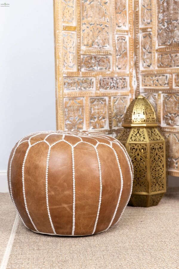 A brown leather ottoman next to a gold lamp.