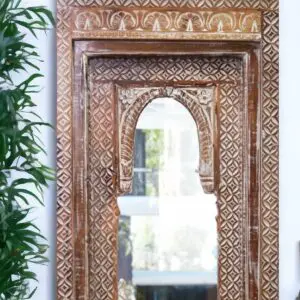 Large temple style mirror. Hand-crafted wooden beveled frame with hand carved detail and arched design, staged in a home next to a plant.