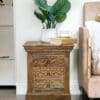 Natural wood nightstand with 3 drawers staged as an accent table in a living room with decorative accents.