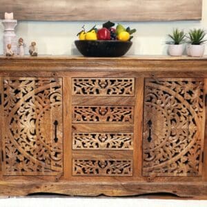 Stunning hand-carved solid natural wood sideboard with 2 carved doors and 3 carved drawers with an intricate cut out design staged in a home with decorative accents.