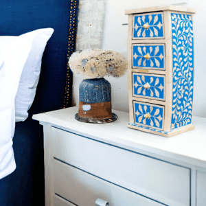 A hand-crafted tall curved jewelry caddy in blue and white bone inlay with a floral patter. Staged on a white nightstand.