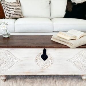 Hand-crafted solid wood white trunk with a dark top and hand-carved detailing. Staged in a living room.
