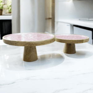 two hand-crafted cake stands, one 12", one 10" in natural wood with light pink enamel tops, staged on a kitchen counter