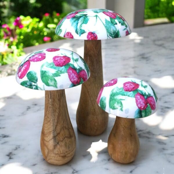 Set of 3 wooden mushrooms with natural wood stalks and enameled caps of white with raspberries and green leaves staged on a marble counter with a garden view in the background