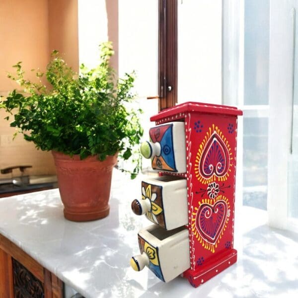 Hand-crafted three drawer organizer with hand-painted detail on the wood exterior, three hand-painted ceramic drawers. Staged in a bight sunny kitchen.