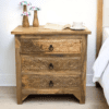 Hand-made from solid mango wood this stunning large 3-drawer side table/nightstand has hand-carved floral drawer fronts. Staged in a sunny bedroom next to a bed.