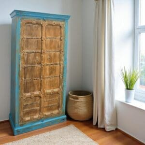 Hand-crafted mango wood armoire in antique style with iron banding and hand-carved detail with blue fame staged in a sunny bedroom