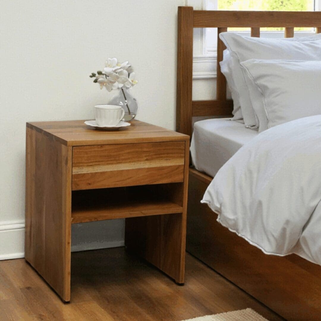 Hand-crafted acacia wood side table with a hidden drawer and shelf staged next to a bed
